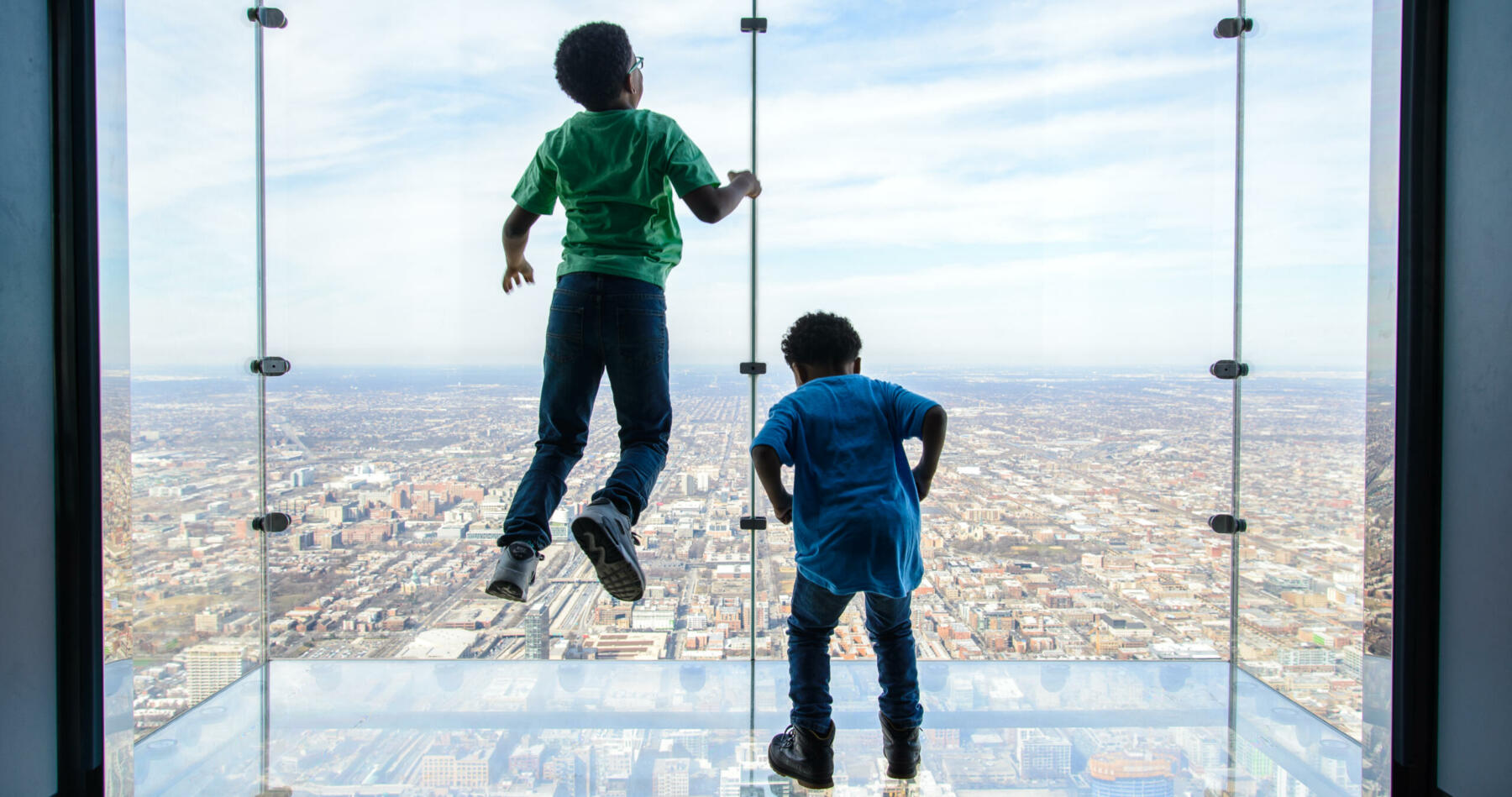 Two children, one standing and one jumping, while taking in the Chicago skyline at the Skydeck.
